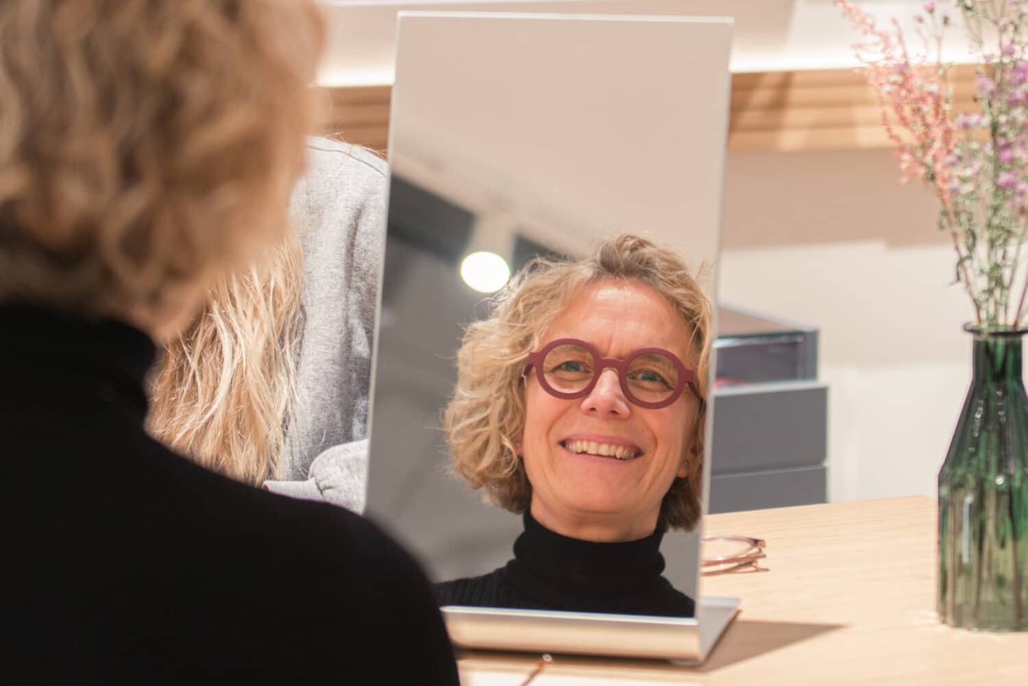 Eine Frau mit blonden Haaren trägt eine rote Brille in einem Brillengeschäft und betrachtet sich lachend im Spiegel.
