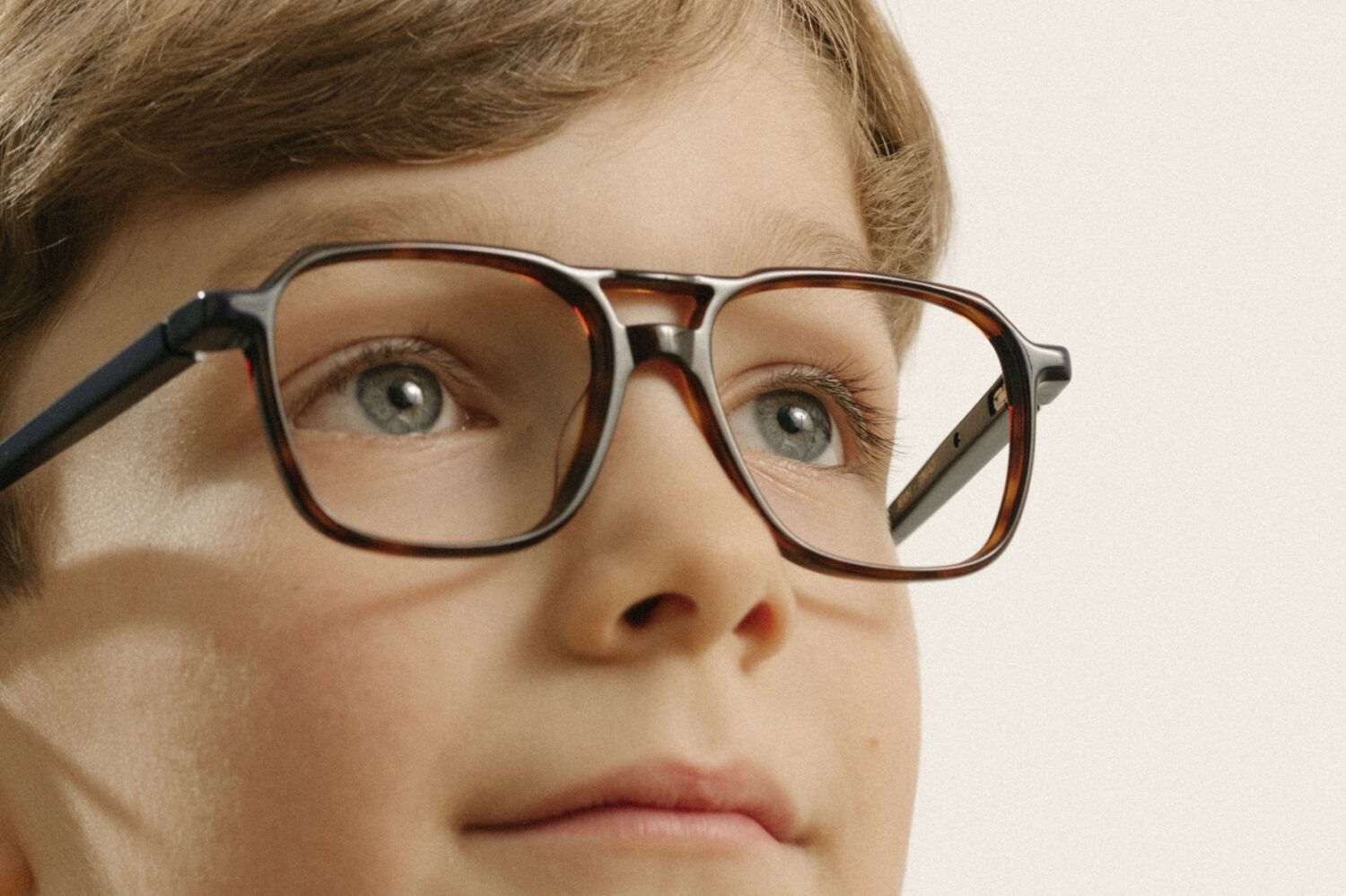 Nahaufnahme eines Jungen mit moderner, dunkel gerahmter Kinderbrille in Braun- und Schwarztönen, der leicht nach oben blickt und seine klaren blauen Augen sowie die gute Passform der neuen Brille zeigt.