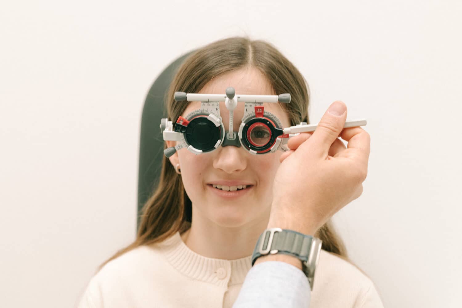 Kind sitzt im Untersuchungsstuhl beim Optiker, trägt eine Messbrille für den Sehtest und blickt nach vorne auf den Prüfbereich, während der Optiker die Werte kontrolliert.