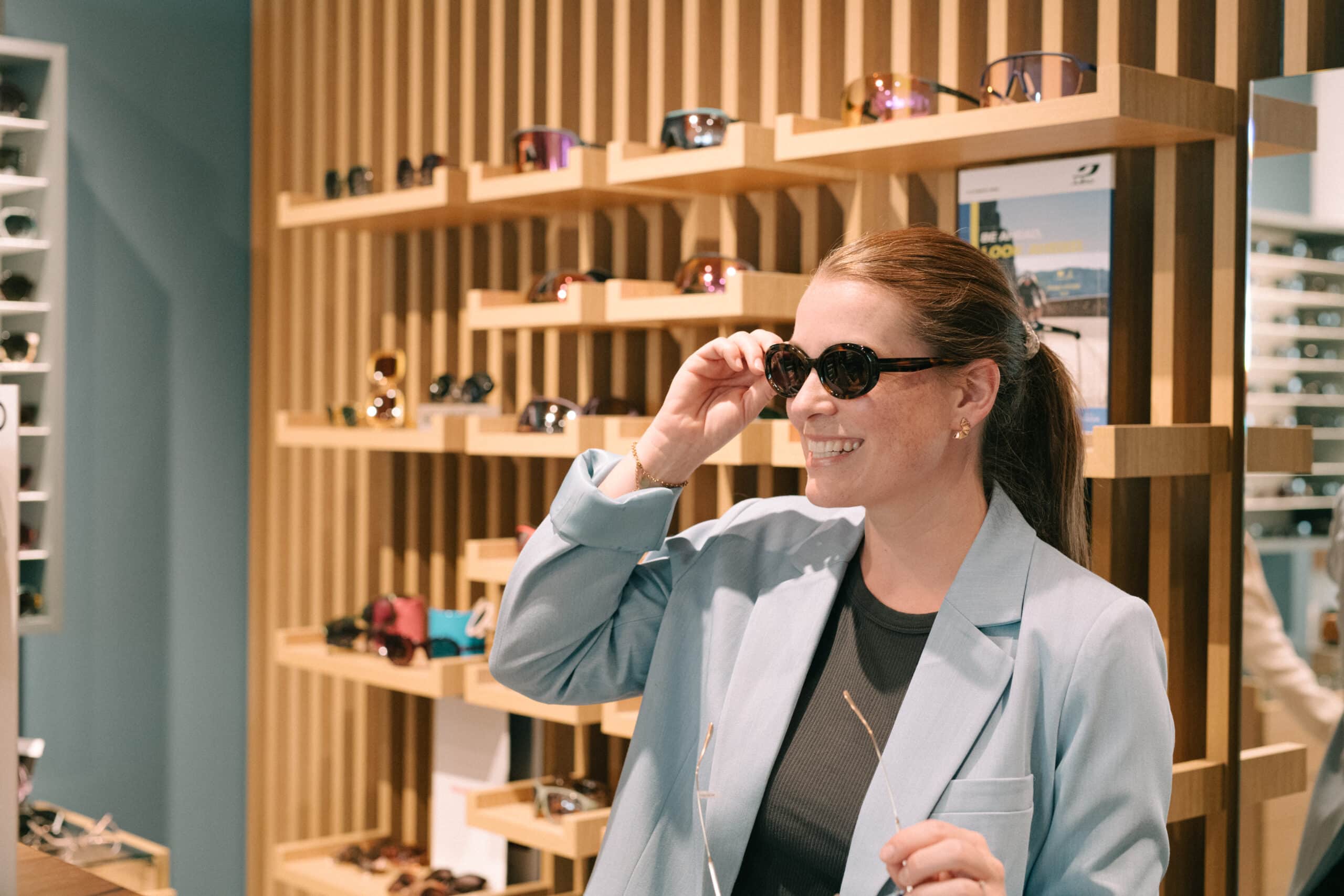 Eine Kundin probiert vor einer modernen Wandverkleidung aus vertikalen Holzlatten eine dunkle Sonnenbrille an. Auf den hölzernen Regalböden im Hintergrund ist eine große Auswahl an Brillenmode ausgestellt.