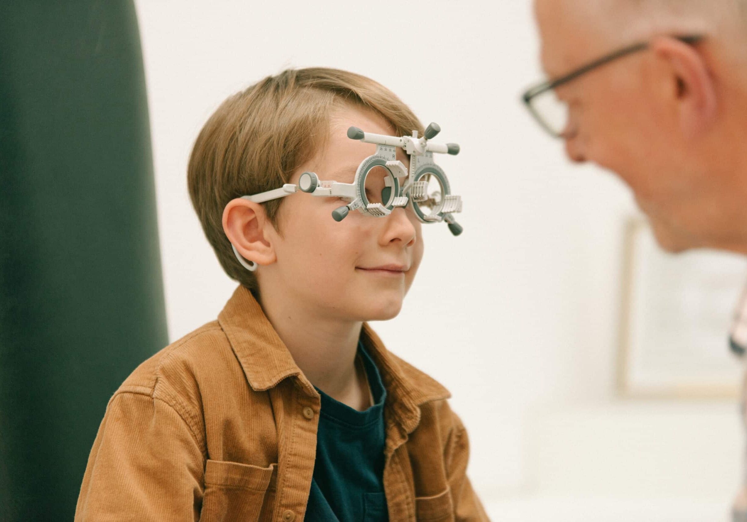 Ein aufgeweckter Junge mit einer ophthalmologischen Messbrille blickt während der Augenprüfung bei Müllers Brillenstudio lächelnd zu seinem Augenoptiker.