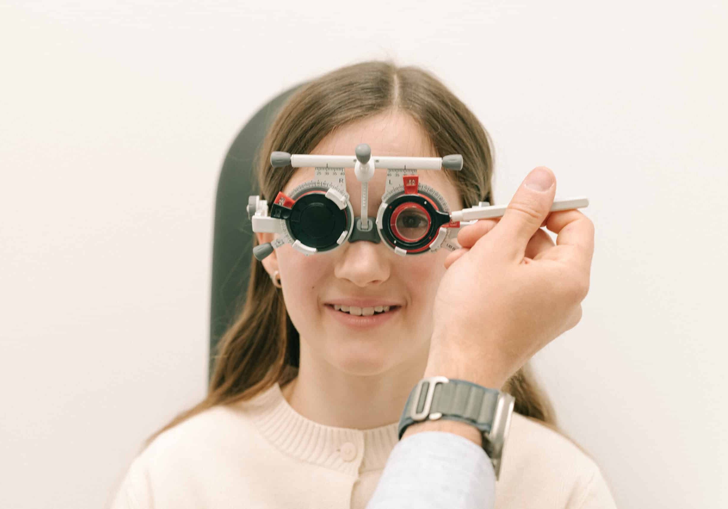 Kind sitzt im Untersuchungsstuhl beim Optiker, trägt eine Messbrille für den Sehtest und blickt nach vorne auf den Prüfbereich, während der Optiker die Werte kontrolliert.