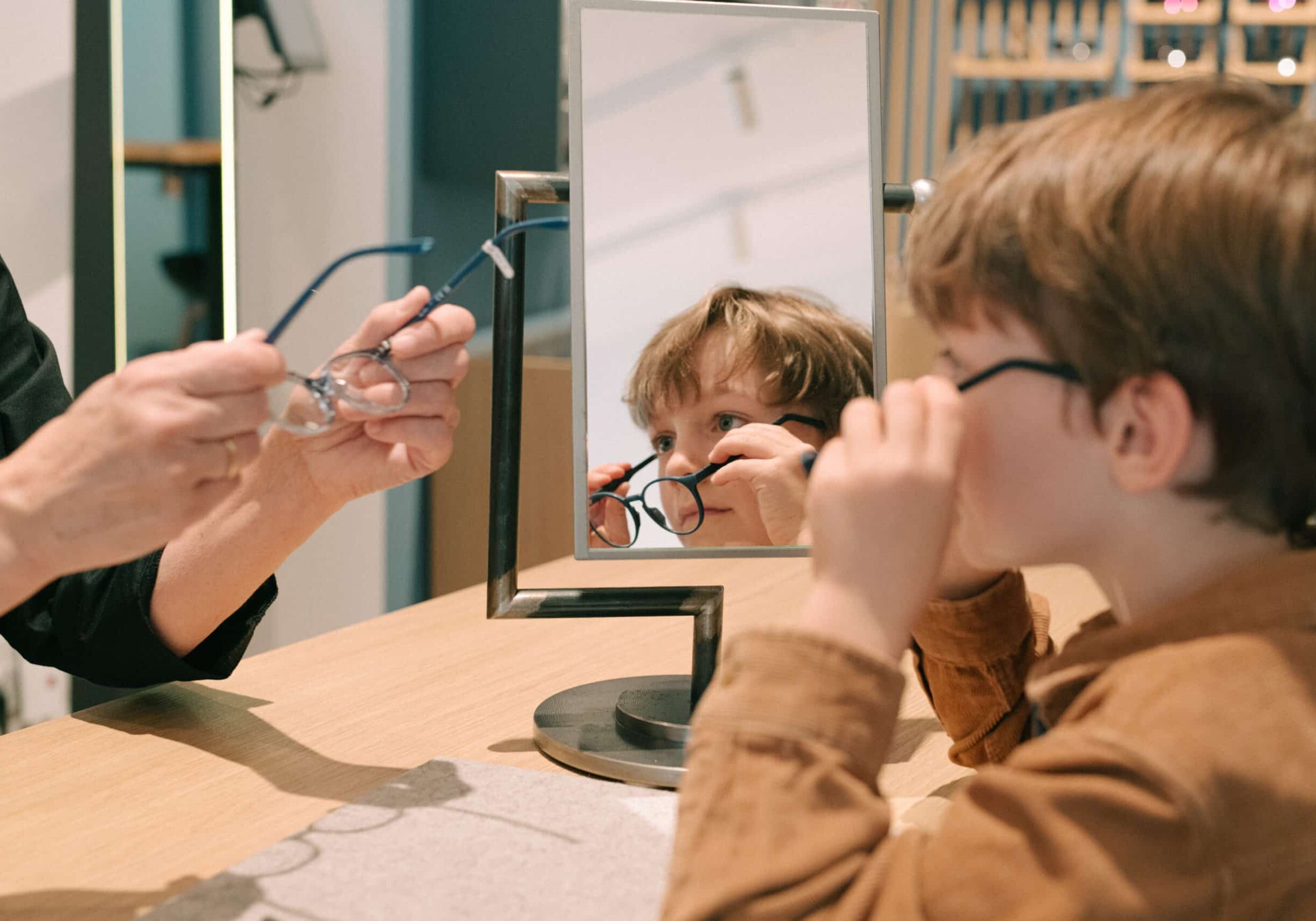 Brillenanpassung für Kinder bei Müllers Brillenstudio. Ein Junge sitzt an einem Holztisch vor einem kleinen Spiegel und probiert eine dunkle Kinderbrille aus, während ein Optiker links ein weiteres Gestell bereithält.