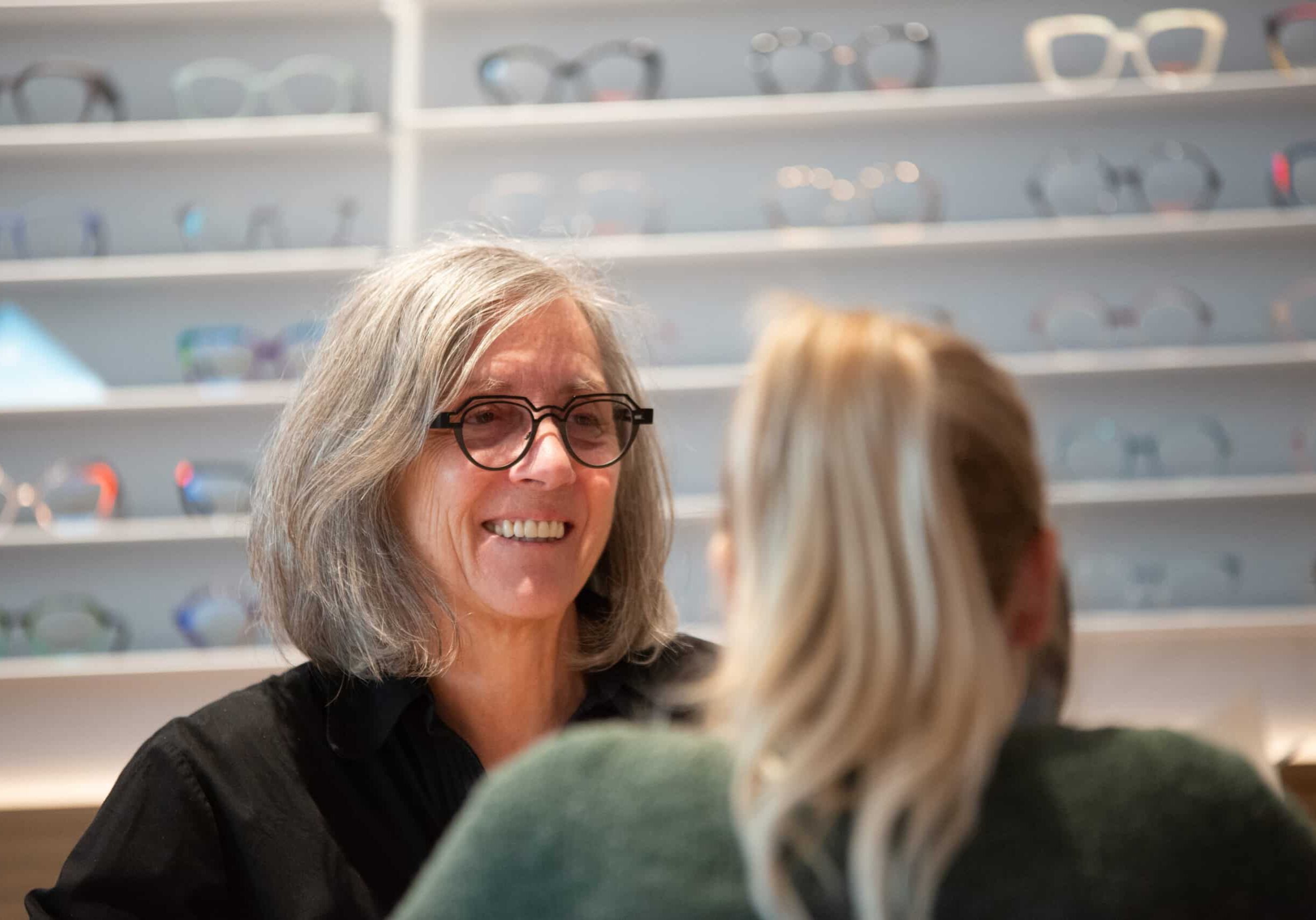 Nahaufnahme einer lächelnden Augenoptikerin von Müllers Brillenstudio mit markanter, dunkler Brille während eines Beratungsgesprächs im Verkaufsraum. Im Hintergrund sind Brillenregale zu sehen.
